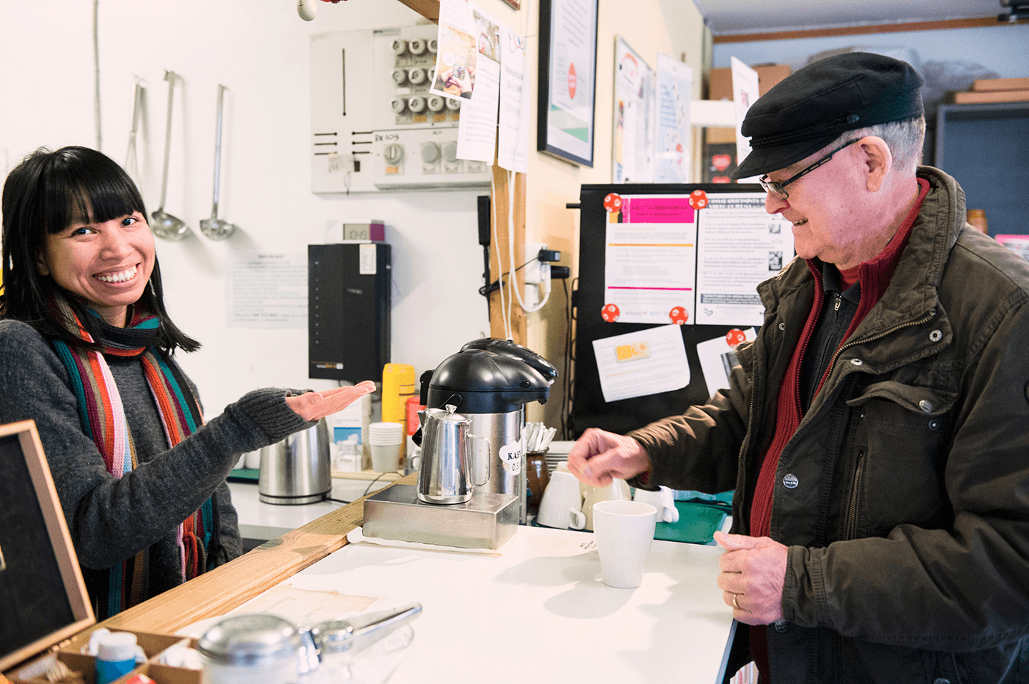 Kuva kohtaamispaikan kahvitiskiltä. Nainen rahastaa ja mies ostaa kahvia.