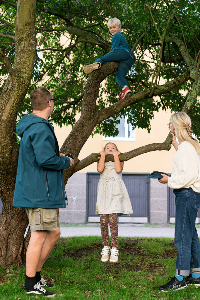 Veli kiipeilee puussa, sisko roikkuu alimmalta oksalta. Isä ja äiti seuraavat vieressä.
