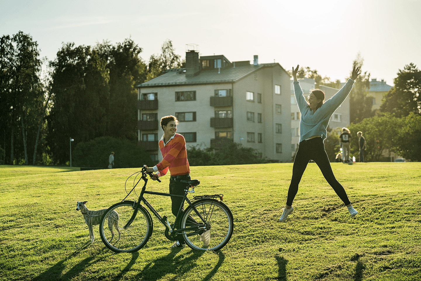 Poika pyöräilee ja taluttaa koiraa kesäisenä päivänä puistossa. Tyttö hyppää ilmaan.