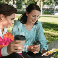 Nuori mies ja nainen lukevat kirjaa ja juovat kahvia puistossa.