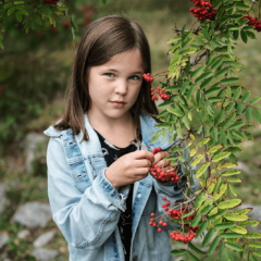 Alakouluikäinen tyttö seisoo metsässä pihlajapuun vieressä.