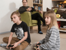 Sisko ja veli pelaa pelikonsolilla ja isä seuraa pelaamista