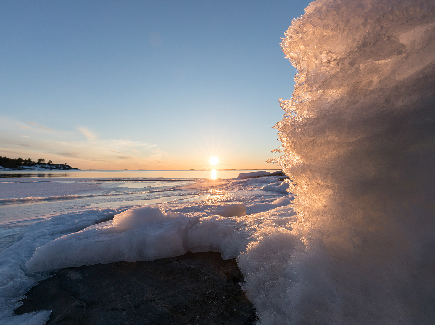 Talvinen maisema, jossa aurinko paistaa matalalla jäätyneen järven taustalla.