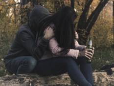 Nainen istuu puunrungolla allapäin, pullo kädess. Toinen ihminen halaa häntä takaapäin.