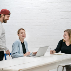 Kolme henkilöä nauraa pöydän ympärillä kannettavan tietokoneen äärellä.