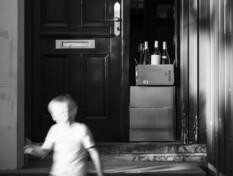 Tekoälyllä tehty mustavalkoinen kuva jossa ovan suussa on postin tuomia viinilaatikoita. Ovelta ulospäin juoksee ylivalaistu lapsen hahmo.